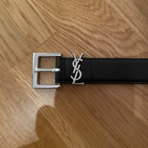 Svart YSL bälte i skinn - Säljer ett stilrent svart bälte från YSL med klassisk silverfärgad logga och spänne. Bältet är smalt och har en enkel, rak form som passar till jeans eller kostymbyxor. Materialet är skinn och detaljerna i metall ger en lyxig känsla.