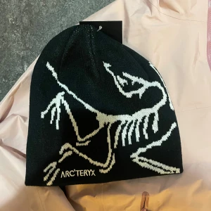 Svart mössa från Arc'teryx - Svart stickad mössa från Arc'teryx med vit dinosaurie-illustration och broderad logga framtill. Snygg och stilren design som passar perfekt för kalla dagar.