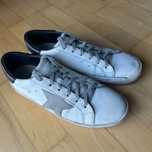 Golden Goose vita sneakers med stjärna - Snygga vita sneakers från Golden Goose med grå mockadetaljer och en ikonisk stjärna på sidan. Skorna har grå snörning, svart häl och platt sula. Perfekt för dig som gillar en cool och avslappnad stil med unika detaljer. Priset är förhandlingsbar.
