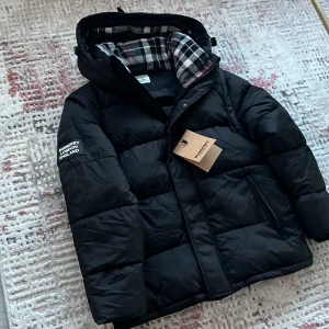 Svart pufferjacka från Burberry - Svart pufferjacka från Burberry med avtagbara ärmar så den kan användas som väst. Jackan har huva med snörning och ett rutigt foder i svart, vitt och rött. Märkespatch på ärmen och dragkedja framtill. Snygg och praktisk för kalla dagar.