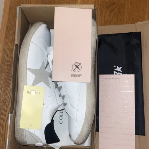 Golden Goose vita sneakers med stjärna - Golden Goose sneakers i vitt skinn med grå mockadetaljer och ikonisk stjärna på sidan. Skorna har grå snörning, svart hälparti och platt sula. Snygg kontrast mellan det vita och gråa, perfekt för dig som gillar en clean men ändå unik look. Storlek 45
