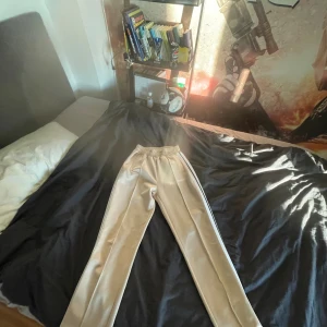 Beige Palm Angels trackpants -  beige byxor från Palm Angels med svart och vit sidostripes. Byxorna har elastisk midja, raka ben och Palm Angels-logga tryckt på framsidan. Qr viden funkar och leder till hemsidan nästan aldrig använt pga för stora