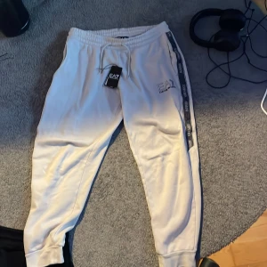 Vita mjukisbyxor EA7 Emporio Armani - Snygga biege mjukisbyxor  från EA7 Emporio Armani med gråa logoband längs sidorna och broderad logga på benet. Byxorna har snörning i midjan och muddar vid bensluten. Perfekta för en chill och sportig look.