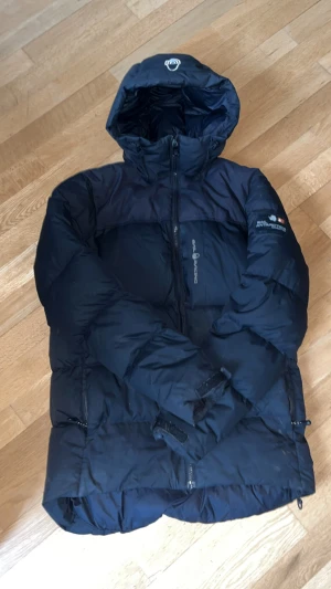 Mörkblå Sail Racing dunjacka - Säljer en mörkblå dunjacka från Sail Racing med huva och snygga logodetaljer på bröstet och ärmen. Jackan har dragkedja framtill, justerbar huva och flera praktiska fickor. Perfekt för kalla dagar och har en clean, sportig look. DET ÄR STORLEK S MEN ÄR LÅNG I STORLEKEN