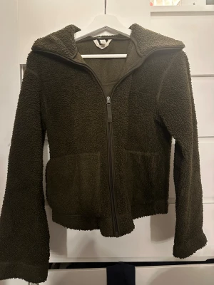 Mörkgrön teddyjacka med huva - Mysig mörkgrön teddyjacka från H&M med huva och två stora fickor framtill. Jackan har en hel dragkedja och är tillverkad i fluffigt fleece-material som håller dig varm under kyliga dagar. Perfekt för dig som gillar chill och comfy stil.