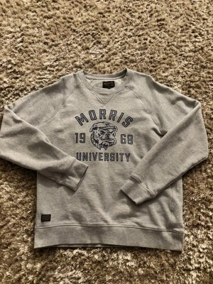Grå sweatshirt Morris University  - Snygg grå sweatshirt från Morris med tryck 'Morris University 1968' och en bulldog på bröstet, storlek M. Tröjan har rund hals, ribbade muddar och är långärmad. Perfekt för dig som vill ha en bekväm och stilren tröja. Jag har knappt använt tröjan då det inte är riktigt min stil. Har du någon fundering så är det bara att skriva👌