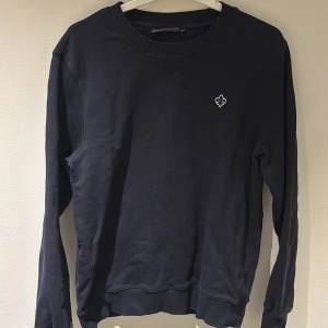 Marinblå sweatshirt från Morris - Marinblå sweatshirt från Morris med rund hals. Tröjan har ett litet broderat emblem på bröstet och är tillverkad i mjuk bomull. En väldigt liten defekt vid höger arm (finns med på bilderna). Hör av dig vid funderingar.