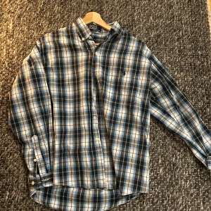 Ralph lauren skjorta - Rutig skjorta från Polo Ralph Lauren i blått, svart och vitt. Klassisk button-down krage, långärmad och med broderad logga på bröstet. Skjortan är tillverkad i 100% bomull och har knäppning framtill.