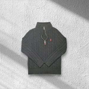 Svart stickad tröja från Polo Ralph Lauren - Svart kabelstickad tröja från Polo Ralph Lauren med röd broderad logga på bröstet. Tröjan har en hög krage med half zip-dragkedja och ribbade muddar vid ärmslut och nederkant. Perfekt för dig som gillar klassisk stil med en modern twist.