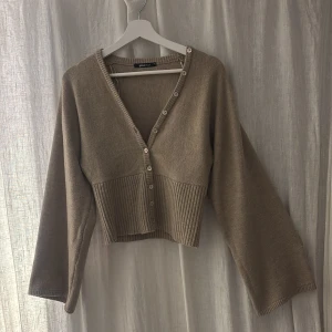 Beige kofta från Gina Tricot XS - Snygg beige kofta från Gina Tricot i storlek XS. Modellen är croppad med bred ribbad nederdel, v-ringad hals och knappar framtill. Koftan har långa ärmar och är stickad i mjukt material som passar perfekt för lager-på-lager. Perfekt för dig som gillar stilrena och enkla plagg.