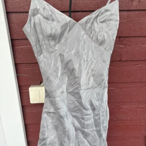 Silver kortklänning från H&M Divided - Snygg silverfärgad kortklänning från H&M Divided i storlek XS. Klänningen har smala axelband, v-ringad hals och skimrande satinliknande material som ger en cool glansig look. Perfekt för dig som gillar minimalistisk stil med lite edge.