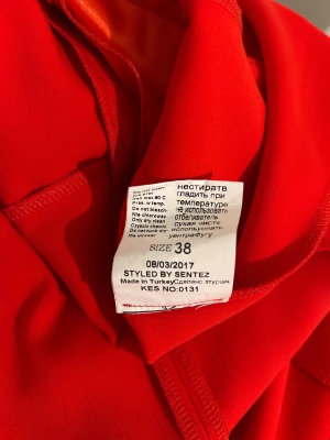 Röd halterneck klänning från Calore - Snygg röd kortklänning från Calore med halterneck och omlott-detalj framtill. Klänningen har en slät yta med glansig insida och dragkedja i ryggen. Passformen är normal och materialet känns som polyester, perfekt för dig som vill sticka ut med en färgstark look.