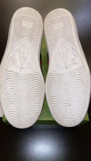 Gucci Ace sneakers med metallic detaljer - Gucci Ace sneakers i vitt skinn med klassiska gröna och röda ränder på sidan. Baksidan har metallicröd och metallicgrön häl med Gucci-logga, och insidan är i mjukt vitt skinn. Snygga snörningar och platt sula. Kommer med originalkartong.