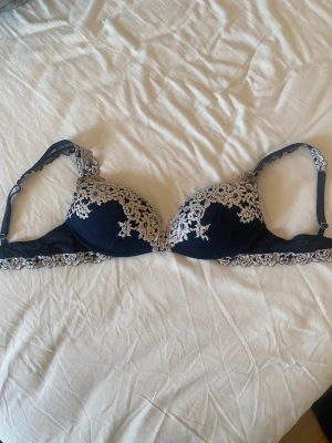 Intimissimi bh - Säljer en mörkblå bh med vacker spets och broderade blommor i vit. Axelbanden är justerbara och har spetsdetaljer, och kanten är dekorerad med broderad spets. Kuporna är vadderade och ger snygg form. Push up från intimissimi. Nästan inte använd köpt för någon vecka sedan, storlek 75b 