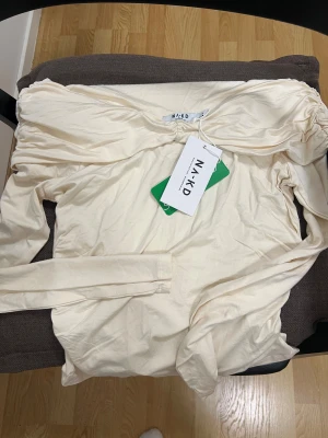 Offshoulder ljusbeige långärmad topp NA-KD - Säljer en snygg offshoulder topp i ljusbeige med lång ärm och tight passform. Toppen har rynkade detaljer längs axlar och sidor som ger en cool och modern look. Materialet är mjukt och stretchigt, perfekt för dig som gillar trendiga plagg med lite extra edge.
