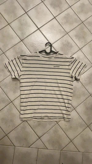 Randig frotté t-shirt från Anerkjendt - Säljer en beige t-shirt från Anerkjendt med svarta horisontella ränder och broderad logga på bröstet. T-shirten är i mjuk frotté och har rund hals samt korta ärmar. Perfekt för dig som gillar chill och stilren vibe.