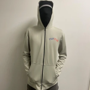 Beige hoodie från Trapstar med tryck - Snygg beige hoodie från Trapstar med färgglatt tryck på bröstet i blått och rött. Tröjan har huva. Passar för stl L