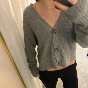 Grå ribbad kofta med knappar - Mysig grå kofta med ribbad struktur och stora, mönstrade knappar framtill. Koftan har en djup v-ringning, långärmade ärmar och en croppad passform som ger en chill vibe. Perfekt att slänga på sig när det är lite kyligt ute.
