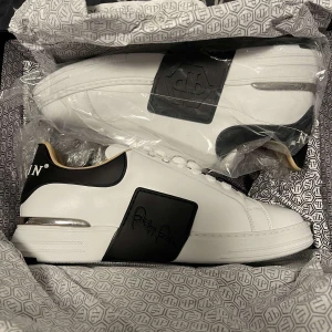 Vita sneakers från Philipp Plein - Snygga vita sneakers från Philipp Plein med svarta detaljer och logotyp. Skorna har snörning och en unik design med en dödskalle på hälen. Perfekta för dig som vill ha en stilren men ändå iögonfallande look.