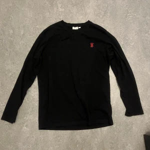 Svart långärmad tröja Burberry - Svart långärmad tröja från Burberry med röd broderad logga på bröstet. Tröjan är tillverkad i 100% bomull och har en klassisk, stilren design med rund hals. Perfekt för dig som gillar enkla och snygga plagg med exklusiv känsla.