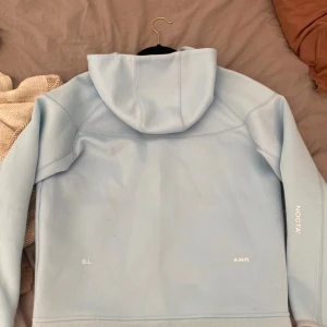 Nike NOCTA ljusblå zip hoodie - Nike NOCTA hoodie i ljusblått med hel dragkedja, huva och två fickor med dragkedja. Snygga vita loggor på bröstet och ärmen. Mjuk och sportig känsla, perfekt för chill eller streetwear. Materialet är bomull och polyester.