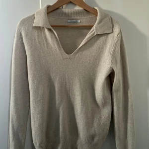 Beige stickad tröja med krage - Snygg beige stickad tröja från Design by sí med klassisk krage och v-ringning. Tröjan har lång ärm och och sitter tajt i armar och lite lösare kring midjan!