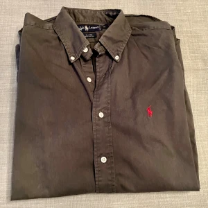 Mörkgrön skjorta från Ralph Lauren - Mörkgrön skjorta från Ralph Lauren med klassisk krage och knappar framtill. Broderad röd Polo-logga på bröstet ger en snygg kontrast. Skjortan är långärmad och oversized, snarare Large än Medium. Se mått på bild.