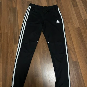 Svarta Adidasbyxor  - Svarta träningsbyxor från Adidas med klassiska vita ränder längs sidorna och Adidas-logga på låret. Byxorna har elastisk midja och är tillverkade i ett mjukt, syntetiskt material som passar perfekt för sport och träning. Osäker på storlek men tror M. Passar S/M
