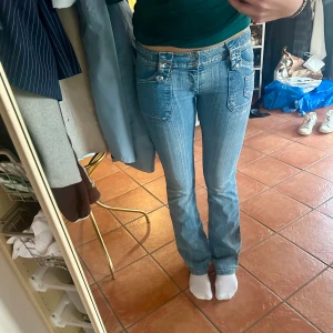 Lågmidjade bootcut jeans - Säljer ett par blå lågmidjade bootcut jeans med låg midja och raka ben. Jeansen har coola detaljer som dubbla knappar, fickor med lock och dekorativa detaljer. Innerbenslängd:83 W:26 Dock väldigt stretchiga 