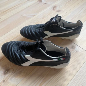 Diadora Brasil Elite fotbollsskor svart/vit 44 1/2 - Säljer ett par Diadora Brasil Elite fotbollsskor i svart och vitt med klassisk design. Skorna har snörning, rund tå och är tillverkade i skinn. Yttersulan har fasta dobbar för gräsplan. Snygg italiensk flaggdetalj på sidan och Diadora-logga på hälen.