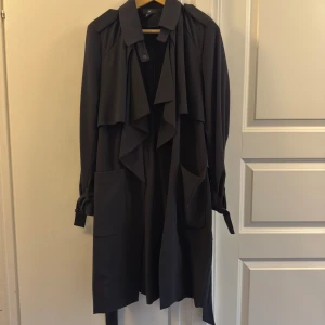 Svart trenchcoat från H&M med bälte - Svart trenchcoat från H&M med snygg vattenfallsframsida, stora fickor och knytband vid ärmsluten. Jackan har bälte i midjan och klassisk krage. Perfekt för lager-på-lager och stilren look under vår och höst. Knälång