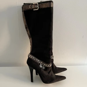 Svarta stövlar med kedjedetalj Rocawear Y2k - Snygga svarta stövlar från Rocawear med spetsig tå och hög klack. Skorna har ett diskret mönster, ormskinnsdetaljer upptill och runt foten samt en silverfärgad kedja med logga. Strl 38 men sitter mer som 37.