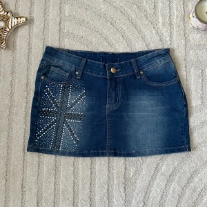 Mörkblå jeanskjol med nitar - Snygg mörkblå jeanskjol med coola nitar och stenar framtill i ett stjärnmönster och detaljer på bakfickan. Klassisk femficksmodell med bälteshällor och knappstängning. Perfekt för dig som vill sticka ut lite extra.