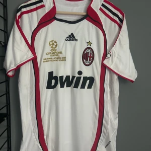 AC Milan Kaka Champions League tröja - AC Milan fotbollströja från Adidas, Champions League-finalen 2007 edition med Kaka och nummer 22 på ryggen. Vit med röda och svarta detaljer, klassisk klubbmärke och Bwin-tryck på bröstet. Tillverkad i lätt och ventilerande Aeroready-material. Helt ny med tagg. En replika