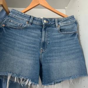 Blå jeansshorts med fransig kant H&M - Snygga blå jeansshorts från H&M i klassisk modell med fem fickor och fransig, rå kant nedtill. Shortsen har normal passform och är perfekta för sommaren. Tillverkade i slitstarkt denimtyg med bälteshällor och knappstängning.