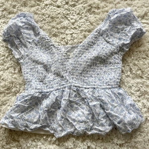 Blommig blus från Hollister XS - Supersöt blus från Hollister i vitt med ljusblått blommönster. Modellen har korta puffärmar, smockad överdel och v-ringning. Volangig nederkant ger en romantisk vibe. Perfekt för vår och sommar, och riktigt skön i bomull.