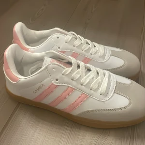 Adidas Samba vit/rosa sneakers - Snygga Adidas Samba sneakers i vitt med ljusrosa detaljer och klassiska tre ränder på sidan. Skorna har vit ovandel i skinn och mocka, gummisula och snörning. 