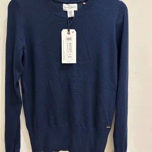 Ny  Marinblå stickad tröja Holly & Whyte - Snygg marinblå stickad tröja från Holly & Whyte med rund halsringning och guldfärgade knappar vid ärmsluten. Tröjan har lång ärm och är i mjukt material som passar perfekt till både jeans och kjol.