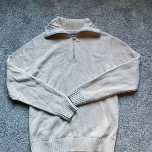 Beige stickad half zip-tröja - Beige stickad tröja från med Jack and Jones ribbad hög krage och half zip-dragkedja. Långärmad modell med breda muddar och klassisk passform. Perfekt för lager-på-lager och chill höst- eller vinterstil. Tröjan är använd och är lite knaprig samt syns lappen genom ett väldigt litet hål i nacken