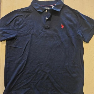 Marinblå piké från U.S. Polo Assn. - Säljer en marinblå pikétröja från U.S. Polo Assn. med klassisk krage och två knappar framtill. Tröjan har en röd broderad logga på bröstet och är tillverkad i mjuk bomull. Perfekt för en stilren och avslappnad look.