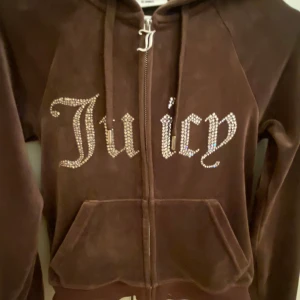 Brun Juicy Couture hoodie i velour - Superfin brun hoodie från Juicy Couture i mjuk velour med glittrande stenar som bildar texten 'Juicy' på bröstet. Tröjan har dragkedja, huva, snörning och fickor framtill. Perfekt för en chill och trendig look. Har används typ 3-4 gånger, säljer den för den e nu tajt för mig storleken passar inte längre mig! Annars bra kvalite, köpt för 650