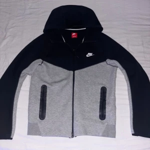 Nike hoodie i svart och grå, M - Kom med pris. Nike hoodie i svart och ljusgrå med hel dragkedja och huva. Snygg tvåfärgad design, vita Nike-loggan på bröstet och svarta detaljer vid fickorna. Perfekt för chill eller träning, mjukt material och långärmad modell.