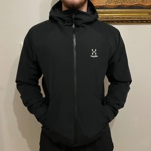 Svart vindjacka från Haglöfs med huva - Svart vindjacka från Haglöfs med minimalistisk design, hel dragkedja och fast huva. Jackan har två sidofickor med dragkedja och diskret logga på bröstet. Tillverkad i lätt och slitstark polyester, perfekt för blåsiga dagar.
