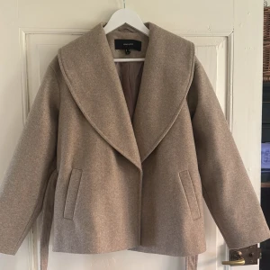 Beige kappa från Vero Moda med bälte - Ny. Stilren beige kappa från Vero Moda med bred krage och knytskärp i midjan. Jackan har raka ärmar, två fickor framtill och är tillverkad i ett mjukt ullblandat tyg. Perfekt för dig som gillar en clean och klassisk look.