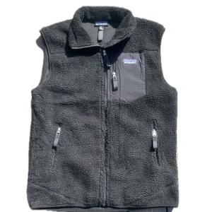 Svart fleeceväst från Patagonia - Mysig svart fleeceväst från Patagonia med hög krage och tre dragkedjeförsedda fickor, varav en på bröstet. Västen har en klassisk, enkel design och är tillverkad i mjuk fleece som håller dig varm. Perfekt att slänga över en hoodie eller långärmad tröja.