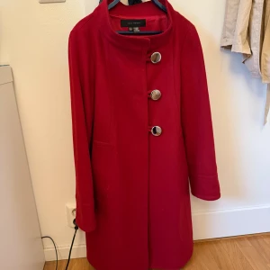 Röd kappa från Zara Woman - Stilren röd kappa från Zara Woman med stora silvriga knappar framtill och dekorativ detalj med knappar i ryggen. Kappan har hög krage, raka ärmar och är tillverkad i ett mjukt ullblandat tyg. Perfekt för dig som vill sticka ut med färg i höst och vinter.