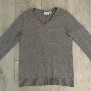 Grå v-ringad stickad tröja från Vila - Mysig grå stickad tröja från Vila med v-ringning och ribbade muddar vid ärmslut och nederkant. Perfekt för lager-på-lager och enkel att matcha med jeans eller kjol. Långärmad och i klassisk modell som funkar året runt. Smått nopprig men inget man tänker på!