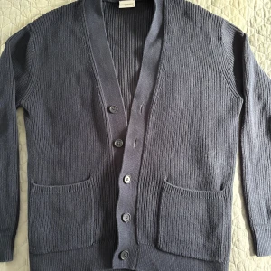 Mörkblå kofta från John Henric - Säljer en stilren mörkgrå kofta från John Henric. Koftan är ribbstickad, har v-ringning, knäppning framtill med fyra knappar och två stora fickor. Perfekt att slänga på sig över en t-shirt eller skjorta för en chill look.