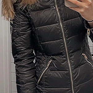 Svart Rockandblue dunjacka med fuskpäls - Snygg svart dunjacka från Rockandblue med guldig dragkedja och detaljer. Jackan har en fluffig fuskpäls på huvan och broderad patch på ärmen. Quiltad design och flera fickor med dragkedja. Perfekt för kalla dagar. Dragkedjan kan gå upp emellan åt! 