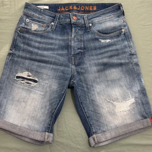 Blå jeansshorts från Jack & Jones - Snygga blå jeansshorts från Jack & Jones med slitna detaljer och uppvikta benslut. Klassisk femficksmodell med knapp och dragkedja i midjan. Shortsen har en cool tvättad look och är perfekta för en avslappnad stil. Märkeslogga på insidan och bak vid linningen.